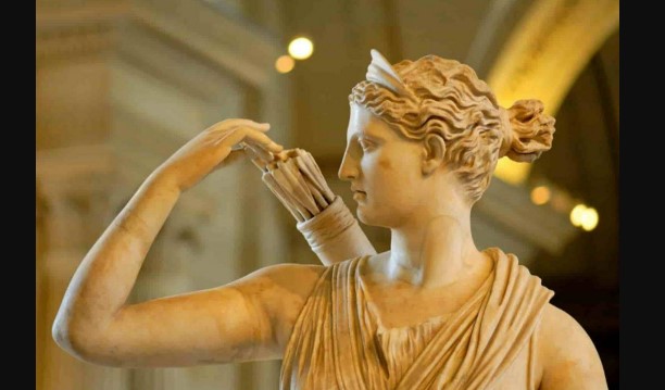 A picture of a statue of Artemis, her curly hair is in a bun and she's reaching back for an arrow