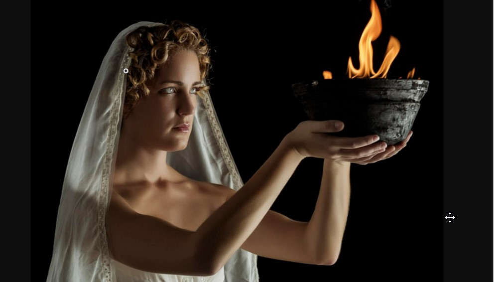 A picture of a model posing as Hesti. She has curly blonde hair and blue eyes, she's wearing a veil and holding a bowl with fire in it.